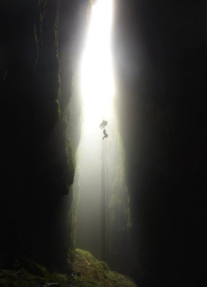 A narrow shaft of daylight falling into a Waitomo tomo, a caver silhouetted on the ropes — adventure caving, not part of the show-cave tours