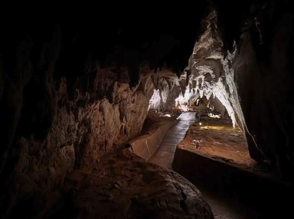 A boardwalk curving through a dark limestone chamber, pale flowstone and stalactites catching the light on the far wall