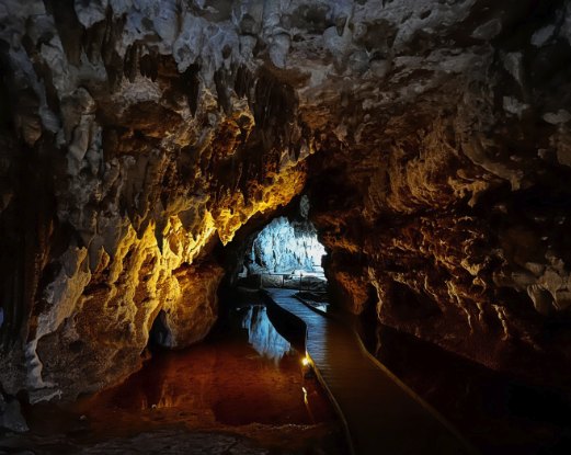 The lit walkway inside the show cave running beside the underground stream, with stalactites overhead and the water reflecting the lamps